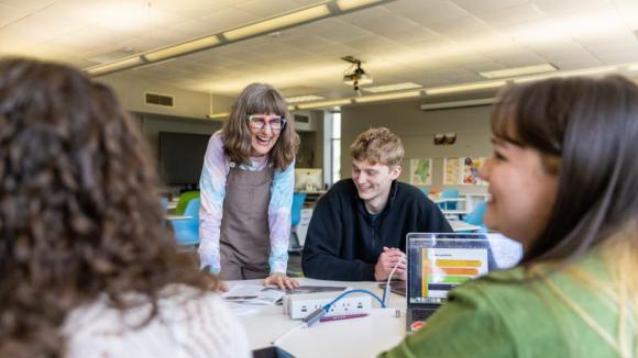 A joyful teacher in colorful glasses interacts warmly with three diverse students around a table in a modern classroom. A laptop shows a bar graph.