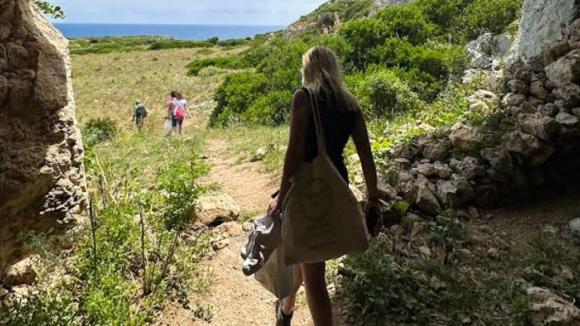 A woman carrying a bag exits a cave onto a sunlit, grassy meadow. Two people walk ahead, with lush greenery and a glimpse of the sea in the distance.