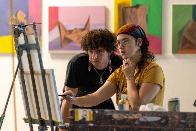Two people focus intently while working on a canvas in an art studio, surrounded by colorful paintings. The atmosphere is creative and thoughtful.