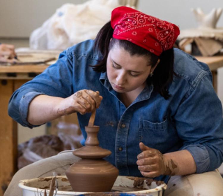 A person in a denim shirt and red bandana focuses on shaping clay pottery on a wheel in a workshop. Crafts and pottery are visible on nearby shelves.