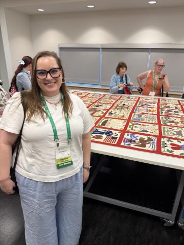 Mary Karcher standing in front of a large quilt spread out on a table with people in the background