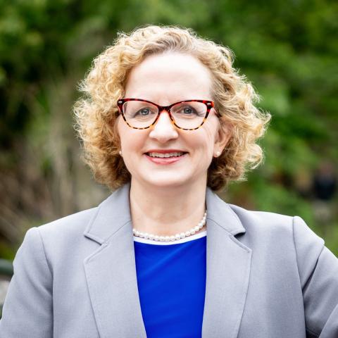 Cathleen Fleck, a white woman with short blonde curly hair and glasses smiling, standing in front of blurred area in a grey blazer with blue shirt, new director