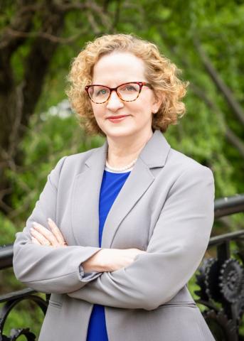 Cathleen Fleck, a white woman with short blonde curly hair and glasses, standing in front of woodsy area in a grey blazer with blue shirt, new director