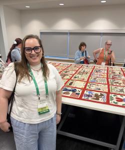 Mary Karcher standing in front of a large quilt spread out on a table with people in the background