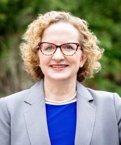 Cathleen Fleck, a white woman with short blonde curly hair and glasses smiling, standing in front of blurred area in a grey blazer with blue shirt, new director