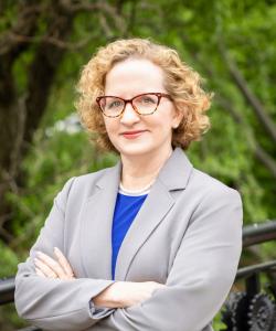 Cathleen Fleck, a white woman with short blonde curly hair and glasses, standing in front of woodsy area in a grey blazer with blue shirt, new director